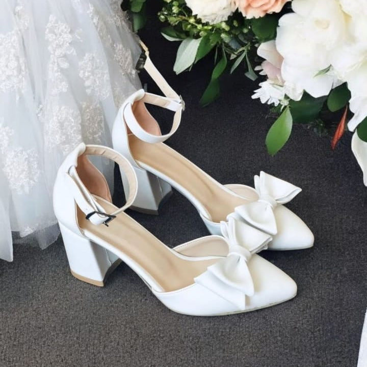 White wedding shoes with bows on a dark surface next to a bouquet of flowers.