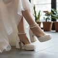 White wedding shoes with pearl straps worn by a person in a white dress.