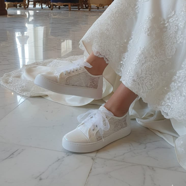 Bride wearing a white lace dress and White lace  sneakers in an elegant room with chandeliers and large windows.